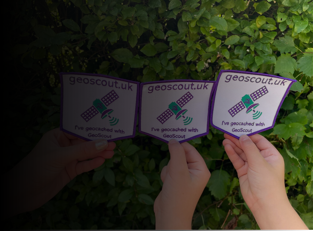 Three GeoScout badges being held up against a green bush